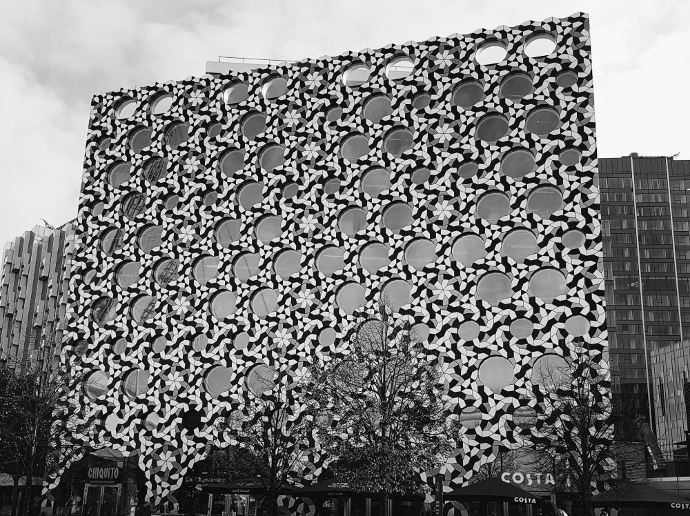 Photograph of the Ravensbourne University building, covered in tiles that together result in a really trippy pattern along with its round windows. That university is really close to The O2 Arena, and which makes getting there by tube or bus extremely easy.