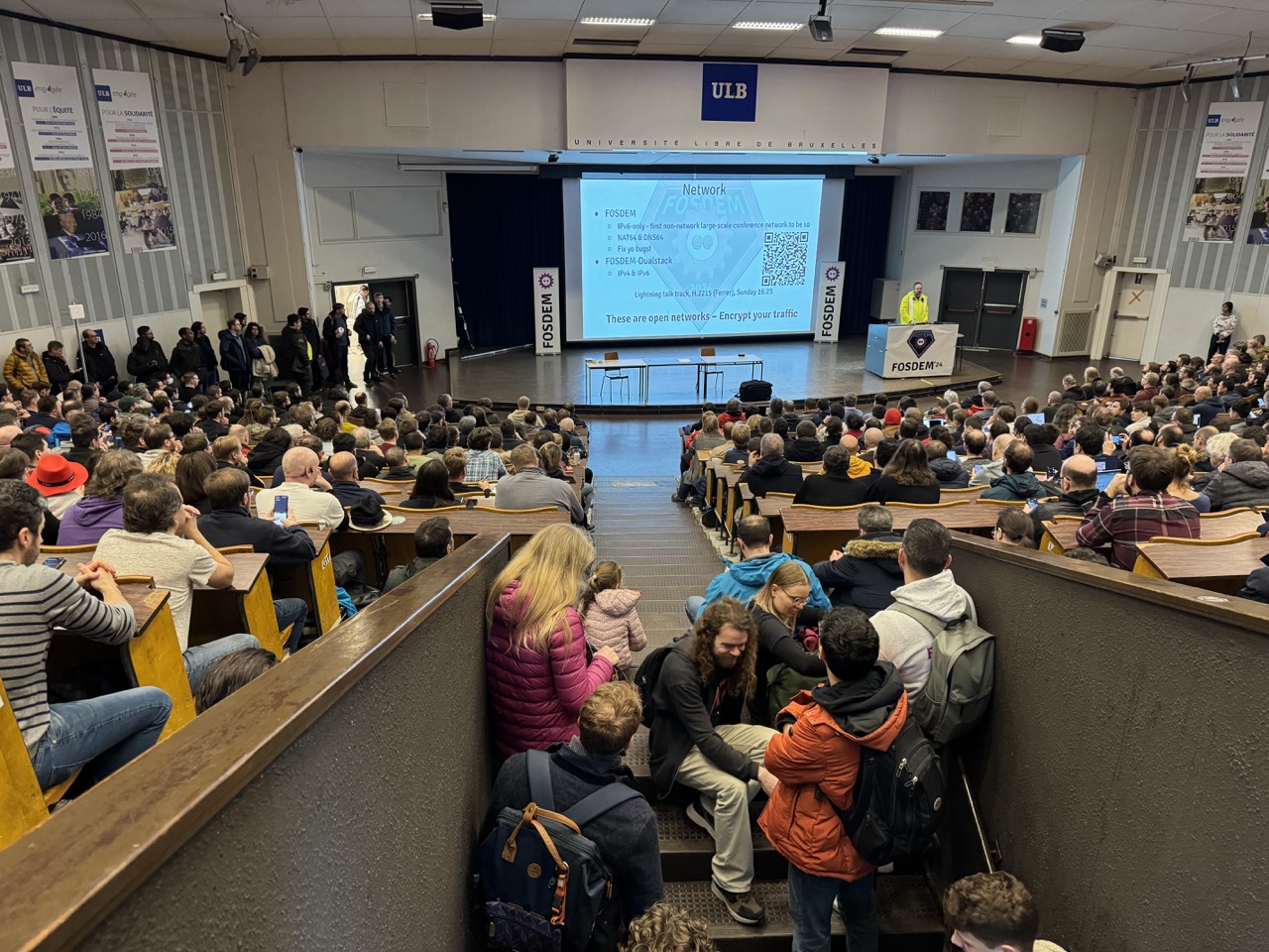 A crowd of people watch one of FOSDEM 2024 organizers talk about this year’s event.